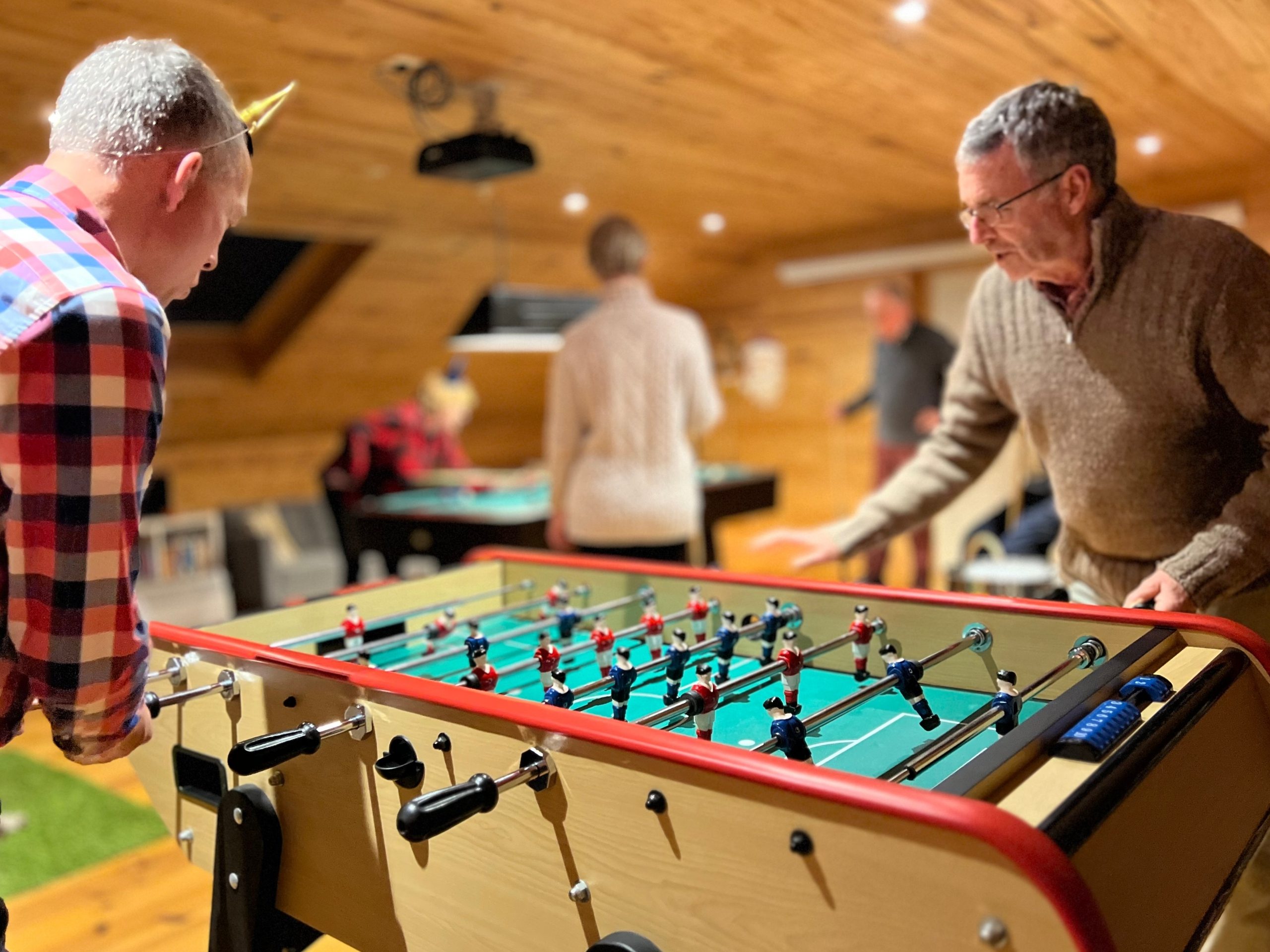 Two men playing Table Football with people playing pool in the background