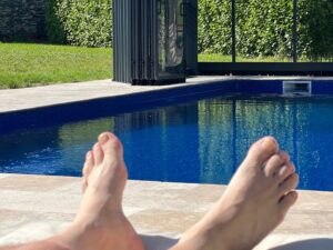 A Pair of mens feet at the end of a sun lounger facing a heated outdoor swimming pool that is deep blue.